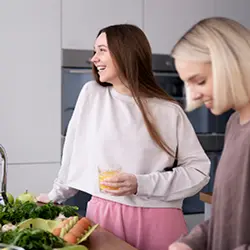 A smiling woman holding a glass of orange juice in a kitchen, standing by a counter with fresh greens and eggs