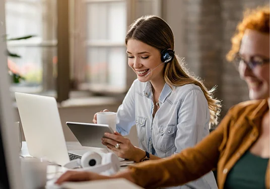 A smiling woman wearing a headset looks at a tablet and holds a mug at a desk, with another woman in the foreground