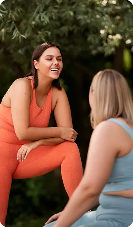 A laughing woman in orange activewear resting her arm on her knee while looking at a woman in blue activewear outdoors