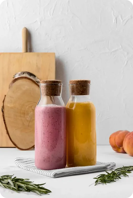 Two cork-topped glass bottles filled with pink and yellow smoothies on a striped cloth, alongside peaches and rosemary